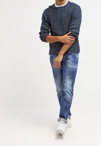 Marinblå stickad tröja med ribbad textur, rund halsringning och långa ärmar, kombinerad med ljusblå slitna jeans och vita sneakers.