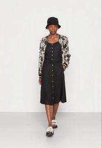 Vestido preto com botões na frente, combinado com uma bomber jacket preta e branca com padrão. A modelo usa um chapéu bucket preto e sandálias brancas.