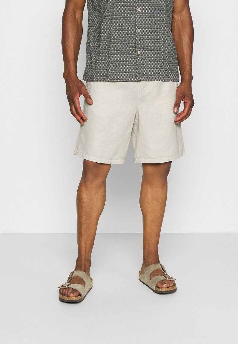 Shorts en lin beige clair, longueur genou, avec une coupe confortable. Portés avec une chemise à manches courtes à motifs foncés et des sandales beiges.