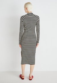 Črna in bela prugasta obleka z dolgimi rokavi, oprijetega kroja, rebraste teksture in stranskim razporkom, v kombinaciji z rdečimi sandalami.