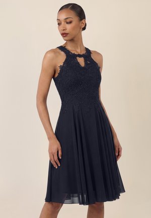 Frau trägt ärmelloses dunkelblaues Kleid mit Spitzenoberteil und Schlitz am Ausschnitt, steht vor schlichtem beigefarbenem Hintergrund.