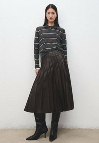 Gonna plissettata marrone scuro con vita alta, abbinata a una camicia a maniche lunghe a righe scure. La modella indossa stivali neri con tacco alto e punta affilata.