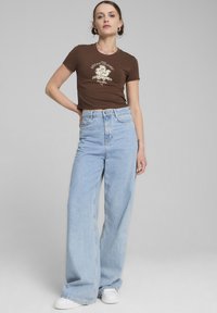 Brun grafisk t-shirt sammen med lyseblå high-waisted vidde jeans. Modellen bærer hvide sneakers. Neutral grå baggrund.