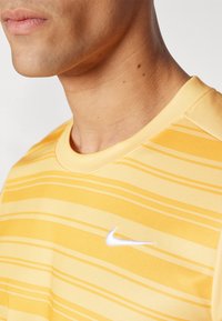 Gul atletisk skjorte med horisontale striper og rund halslinning. Den har en liten hvit Nike-logo på venstre side. Glatt tekstur.