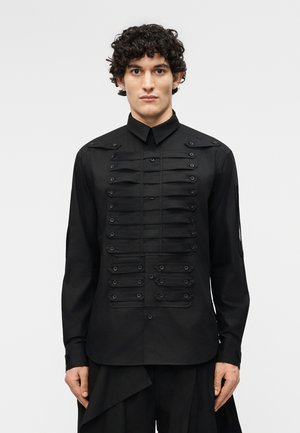 Νέος άνδρας με σγουρά μαλλιά που φοράει μαύρο πουκάμισο με κουμπιά, διακοσμημένο με οριζόντιες λουρίδες και κουμπιά στο μπροστινό μέρος και στα μανίκια.