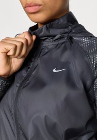 Zwarte windbreaker van Nike met een gladde, glanzende textuur, een ritskraag en reflecterende stippenaccenten op de mouwen. Logo aan de voorkant geplaatst.