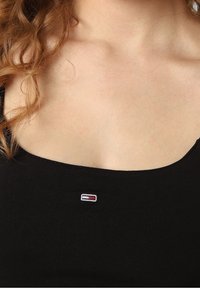 Czarna bezrękawnik z dekoltem w serek, z małym haftowanym logo w czerwonym, białym i niebieskim na części piersiowej. Gładka tekstura.