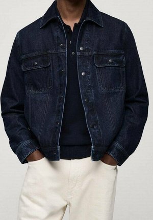Mand iført mørkeblå denimjakke over sort skjorte og off-white bukser, hænder i lommerne, overkrop synlig mod neutral baggrund.
