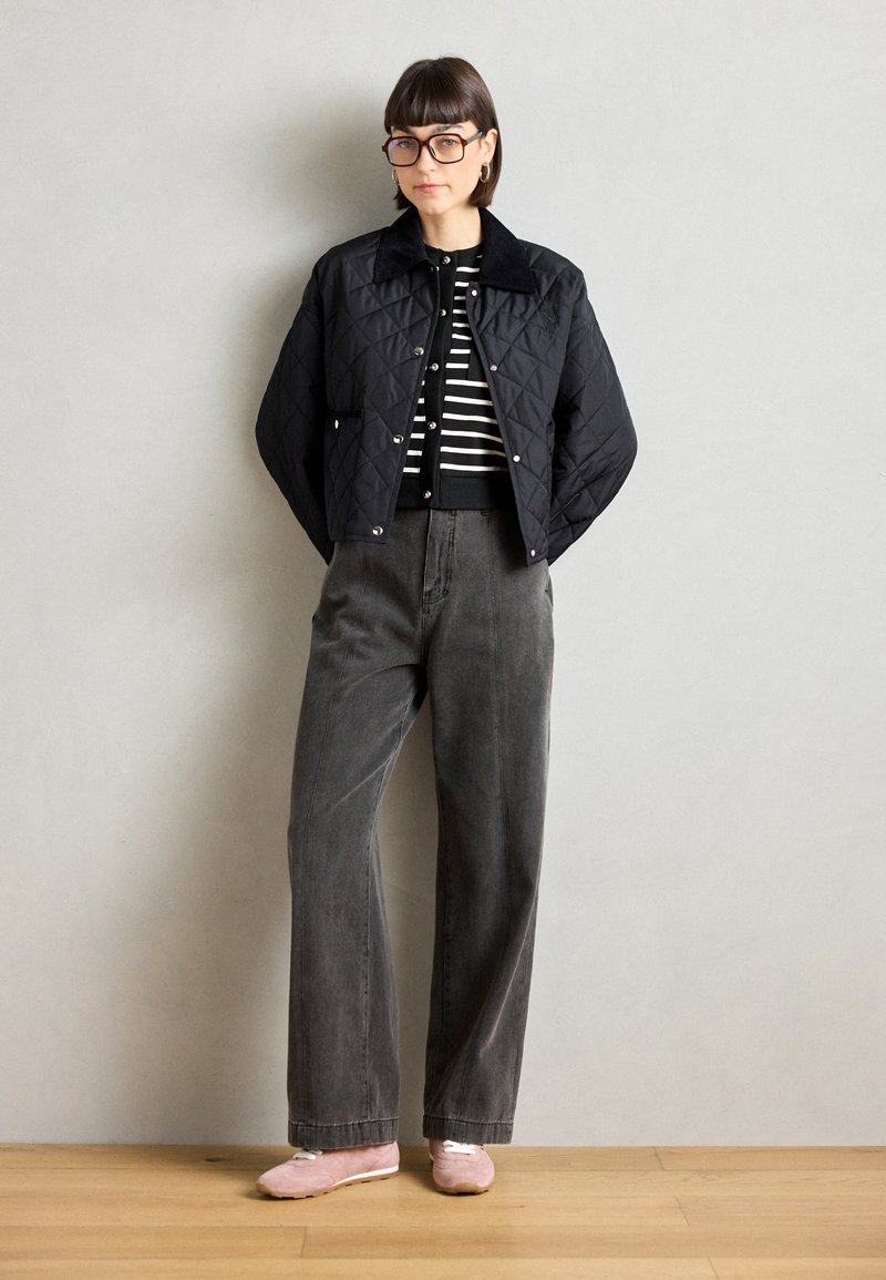 Zwarte gewatteerde jas met drukknoopjes, gedragen over een zwart-wit gestreept shirt, gecombineerd met grijze brede broek en rozige instapschoenen.