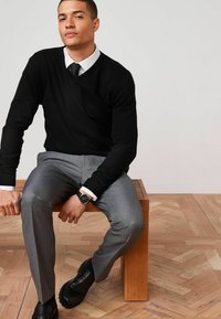 Suéter negro de cuello en V, camisa blanca, pantalones de vestir grises, zapatos negros y reloj. Modelo sentado en un banco de madera con un suelo texturizado.