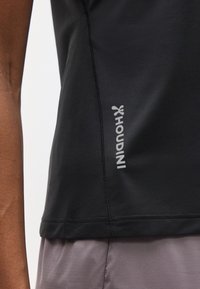 Κοντινό πλάνο ενός ατόμου που φοράει ένα μαύρο αθλητικό μπλουζάκι με το σήμα Houdini και εμφανείς ραφές πάνω από ανοιχτό γκρι παντελόνια.