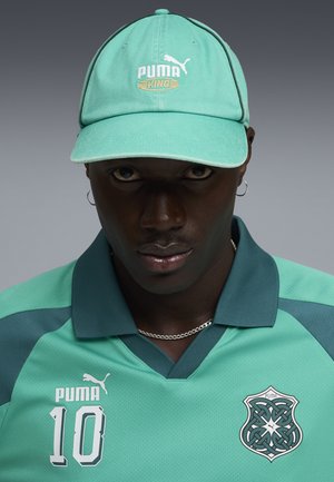 Hombre con gorra turquesa de Puma y camiseta deportiva a juego con el número 10 y el logo de Puma estilo celta, sobre fondo gris.