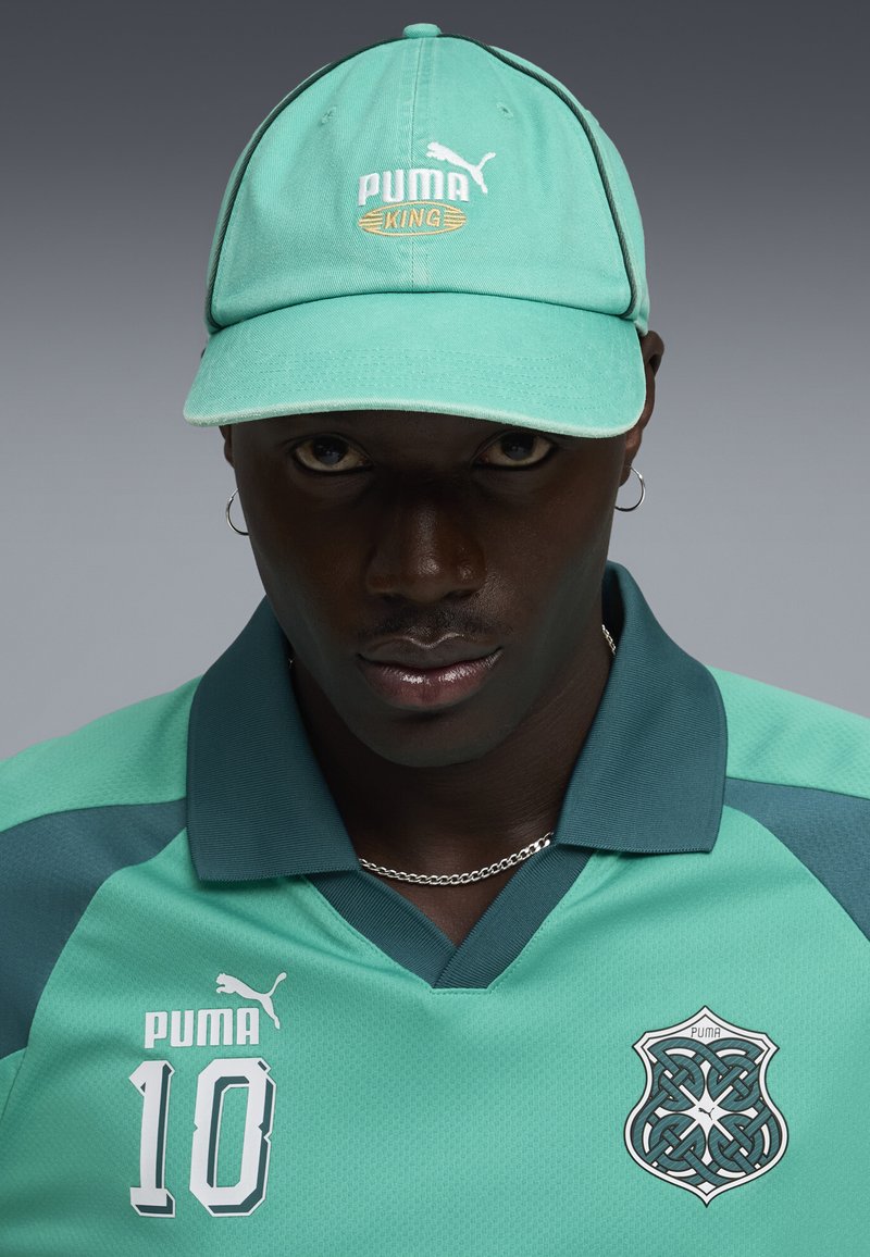 Homme portant une casquette Puma turquoise et un maillot de sport assorti avec le numéro 10 et un logo Puma de style celtique, sur fond gris.