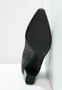 Chaussure noire à talon haut avec une tige texturée, un bout pointu et une semelle à motif. Le talon a une légère courbure et une finition mate.