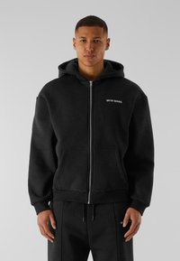 Haut à capuche noir zippé avec une poche avant, fabriqué en tissu doux. Il possède une capuche avec cordon de serrage et un petit logo sur la poitrine.
