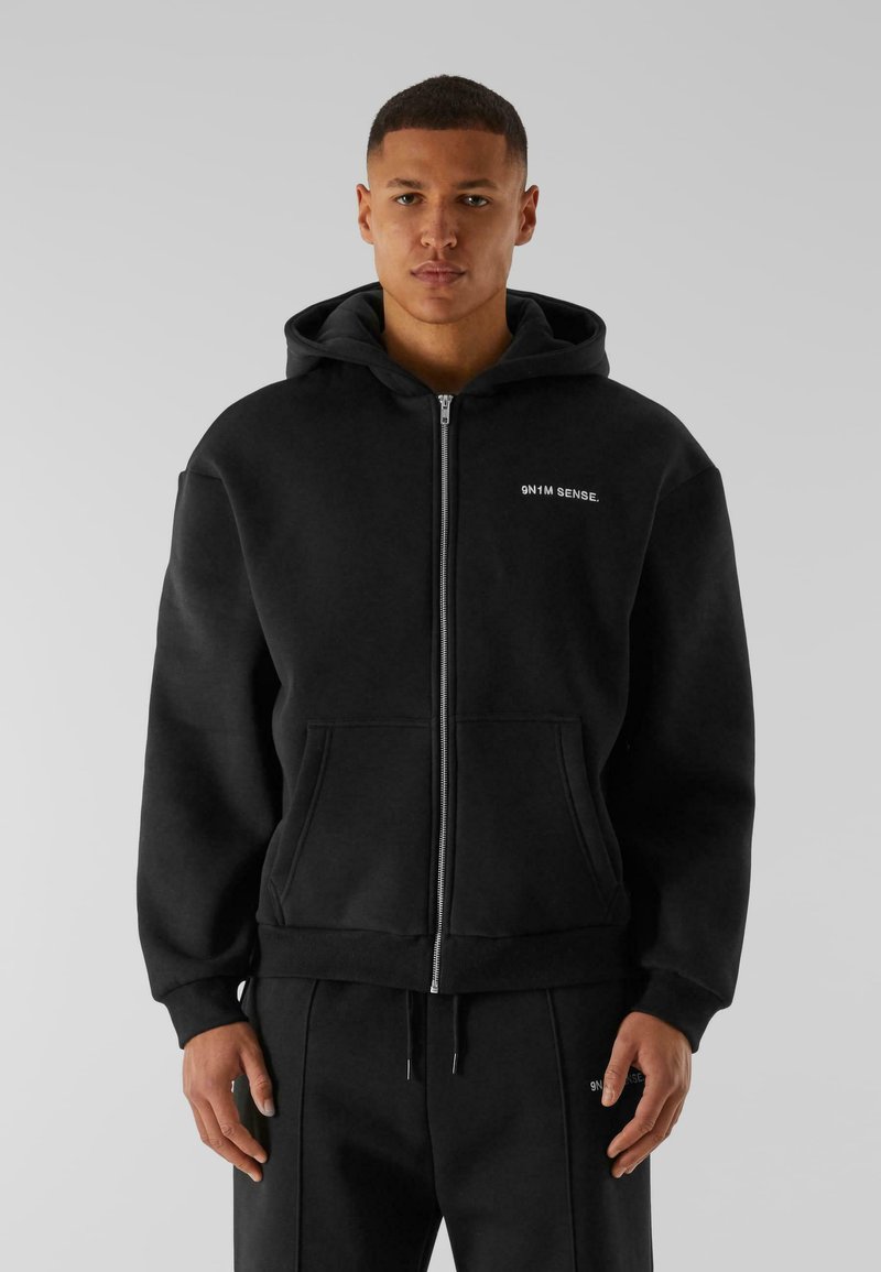 Haut à capuche noir zippé avec une poche avant, fabriqué en tissu doux. Il possède une capuche avec cordon de serrage et un petit logo sur la poitrine.