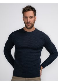 Pulover texturat de culoare bleumarin, cu guler rotund și manșete răsucite, având un model țesut și un logo mic pe tiv.