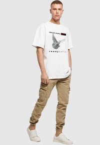 Hvid oversized t-shirt med en grafik af en due, teksten "upscale studios SKY" og "ready to fly". Sammen med beige cargo bukser og hvide sneakers.