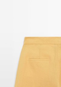 Vue arrière d'une jupe jaune moutarde montrant la taille, une couture verticale et une poche passepoilée unique sur fond blanc uni.