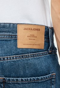 Καφέ δερμάτινο έμπλαστρο σε μπλε τζιν με κείμενο «JACK & JONES, Jeans Intelligence, Since 1990, Craftsmanship and Dedication» κοντά στη μέση.