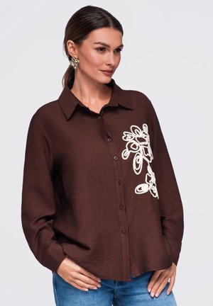 Femme portant une chemise marron à boutons avec une broderie florale blanche sur le côté gauche, un jean bleu et de grandes boucles d'oreilles dorées, posant les mains dans les poches.