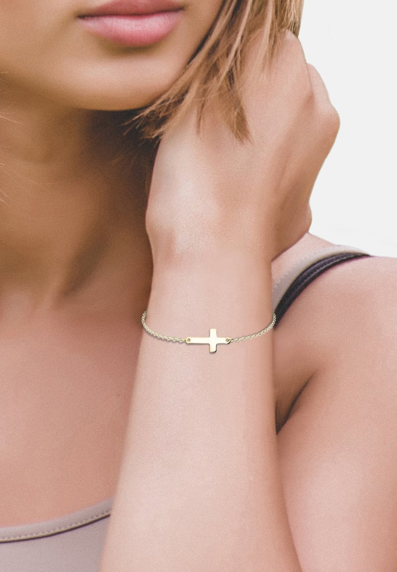 Goldfarbener Armreif mit einem minimalistischen Kreuzanhänger, ausgestattet mit einer zarten Kette und glatter Textur, am Handgelenk vor einem hellen Hintergrund getragen.