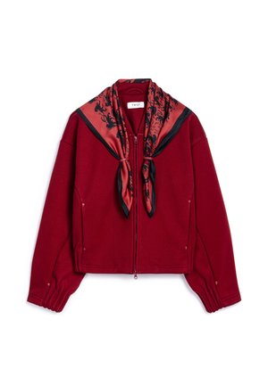 Veste bordeaux raccourcie avec des manches longues, fermeture éclair et une écharpe rouge avec des motifs floraux noirs nouée au col.