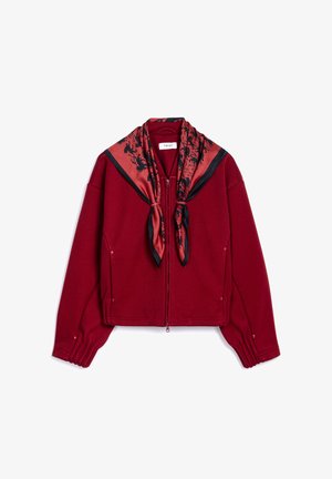 Veste bordeaux raccourcie avec des manches longues, fermeture éclair et une écharpe rouge avec des motifs floraux noirs nouée au col.