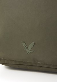Bolsa de tela verde con una superficie texturizada que presenta un logo de águila en relieve. Diseño simple y rectangular con detalles cosidos.