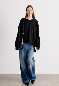 Sweatshirt nero oversize con spalle abbassate, abbinato a jeans in denim blu a gamba larga e scarpe nere. Piccola borsa nera con tracolla.