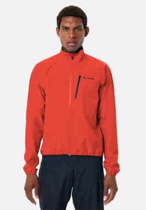 Uomo che indossa una giacca rossa con cerniera e tasca sul petto e pantaloni neri, in piedi davanti a uno sfondo chiaro e uniforme.