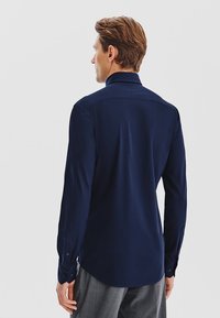 Granatowa koszula z długim rękawem z klasycznym kołnierzykiem, z zapięciem na guziki przy mankietach oraz gładką teksturą. Posiada zaokrąglony dół i dopasowany krój.