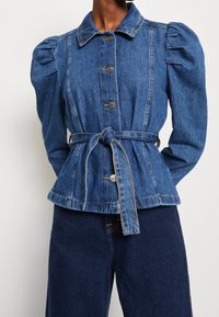 Giacca di jeans con maniche a sbuffo, chiusura a cinque bottoni e vita cintrata con cintura. Presenta un colletto e un orlo sfrangiato. Colore blu intenso.