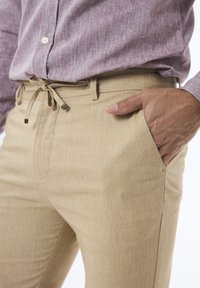 Pantalons beige avec une finition texturée, présentant une taille à cordon de serrage et des poches latérales, associés à une chemise à boutons légère de couleur mauve.