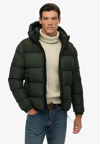 Mørkegrøn pufferjakke med hætte, quiltet design, lynlåslommer, båret over en cremefarvet strikket turtleneck og blå jeans. Tekstureret stof.