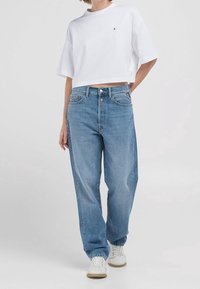 Kvinna i en vit kortärmad T-shirt, avslappnade blå jeans och vita sneakers, stående med en hand bakom ryggen.