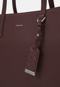 Calvin Klein vinröd tote-väska i texturerat syntetmaterial, med en silverfärgad logotyp och en tagg med ett upprepat CK-mönster.