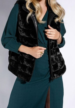 Frau trägt ein türkisfarbenes Kleid mit einem Vorderseitenschlitz und eine ärmellose schwarze Kunstfellweste, accessoriert mit goldenen Ringen und Armbändern.