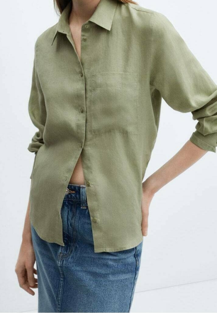 Personne portant une chemise vert clair boutonnée avec les manches retroussées et un jean bleu taille haute, debout avec une main sur la hanche.