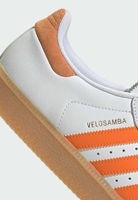 Białe sneakersy z pomarańczowymi zamszowymi akcentami, teksturowaną skórą, równoległymi pomarańczowymi paskami oraz gumową podeszwą w zygzakowaty wzór. Nazwa marki "VELOSAMBA" widoczna.