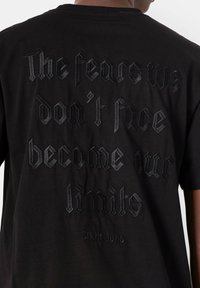 Zwarte katoenen T-shirt met reliëftekst in een Gotische schreefletter op de rug, met de tekst "De angsten die we niet onder ogen zien, worden onze grenzen."