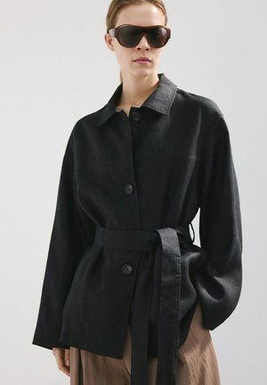 Žena v nosenej veľkej čiernej bunde so zipsom a pásom a veľkými tmavými slnečnými okuliarmi na jednoduchom svetlom pozadí.