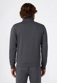 Uomo con capelli ricci che indossa una felpa grigia scura a maniche lunghe e pantaloni abbinati, visto di spalle su uno sfondo bianco uniforme.
