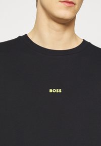 Svart bomullströja med rund halsringning, med en kontrasterande gul "BOSS"-logotyp centrerad på bröstet. Enkelt och avslappnat design.