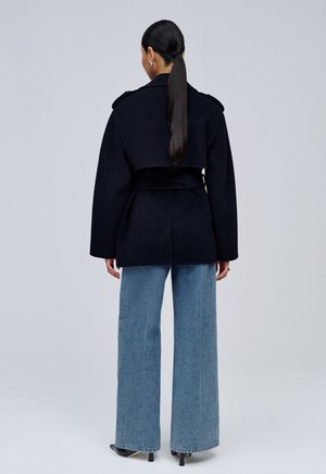 Femme avec une longue queue de cheval foncée, portant un manteau noir ceinturé, un jean bleu à jambes larges et des chaussures noires à talons, debout de dos sur fond gris.