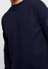 Marinblå långärmad skjorta i texturerad tyg, med en liten svart logotyp på bröstet och en subtil design på ärmarna.