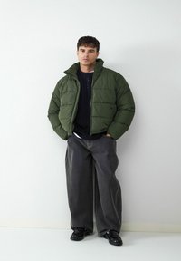 Grüne Pufferjacke mit hohem Kragen, gerippter Textur und gesteppten Paneelen. Kombiniert mit dunkelgrauen weit geschnittenen Hosen und schwarzen Schuhen.
