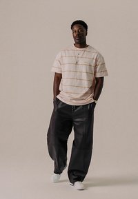 Ροζ ριγέ μπλουζάκι με άνετη γραμμή, σε συνδυασμό με φαρδιά μαύρα δερμάτινα παντελόνια και λευκά αθλητικά παπούτσια, στέκεται μπροστά σε ουδέτερο φόντο.