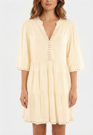 Mujer con un vestido suelto de color crema, con mangas tres cuartos y detalles de tachuelas plateadas en el escote, los puños y el dobladillo.