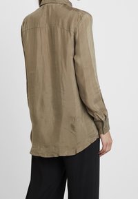 Olivově zelená košile s dlouhým rukávem vyrobená z hladké, lehké látky. Má límeček a zapínání na knoflíky u manžet, s volným střihem a zakřiveným lemem.